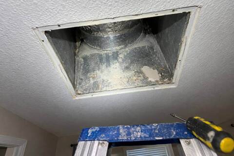 Ceiling vent opening with its grille removed, pale dust and a scrap of drywall resting on the metal inside, seen from a stepladder