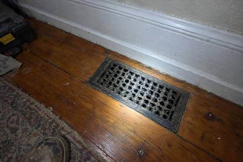 Cast-iron floor register set into worn pine floorboards next to a painted baseboard, lit by a flashlight