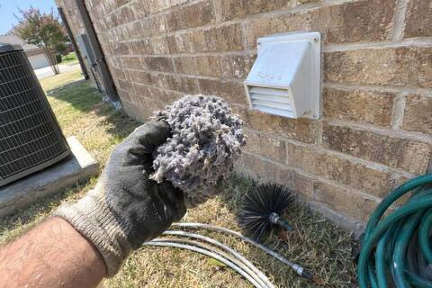 Gloved hand holding a thick clump of gray lint in front of a dryer vent hood on a brick wall, brush rods lying on the grass