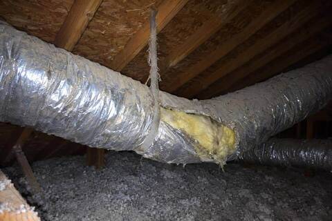 Silver flexible duct sagging between attic rafters, its outer jacket torn open and yellow insulation showing, held up by one strap