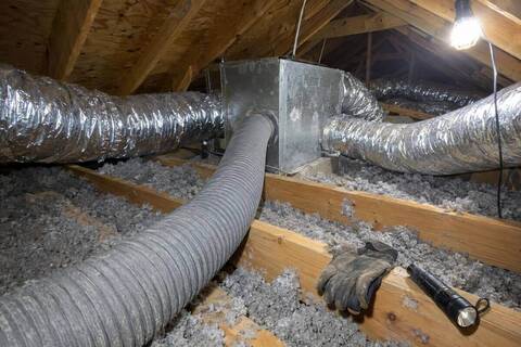 Wide ribbed vacuum hose lying across attic joists toward a sheet-metal box, with silver insulated ducts branching off over loose insulation