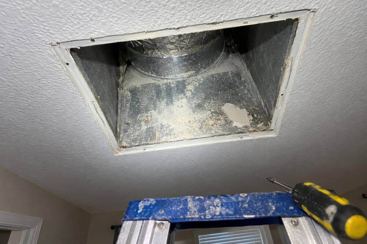 Ceiling vent opening with its grille removed, pale dust and a scrap of drywall resting on the metal inside, seen from a stepladder