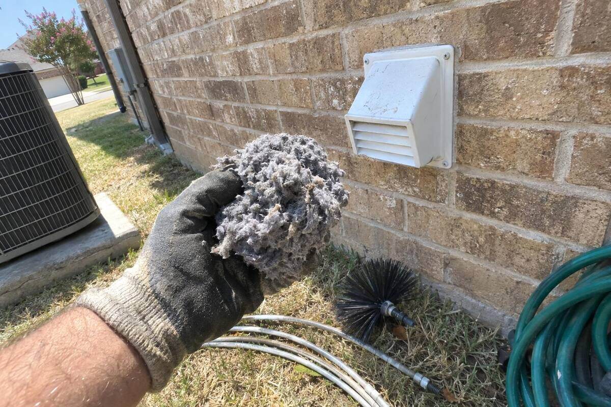 Gloved hand holding a thick clump of gray lint in front of a dryer vent hood on a brick wall, brush rods lying on the grass